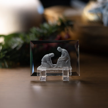 A clear rectangular crystal plaque on a small clear stand, featuring a white relief sculpture of Jesus cleaning his apostles feet and the inscription 'LUKE 22:27' on the front, displayed on a wooden surface with a softly blurred indoor background.