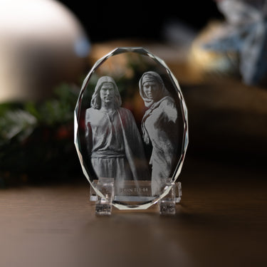 A clear acrylic or glass oval plaque on a small clear stand, featuring a grayscale photo ofJesus Christ and Mary at the tomb of Lazarus and the engraved text 'JOHN 11:1-44' at the bottom, displayed on a wooden surface with a blurred indoor background.