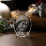 A clear oval crystal plaque featuring an engraved portrait of Jesus Christ, displayed on a small clear stand on a wooden surface with a softly blurred holiday background.