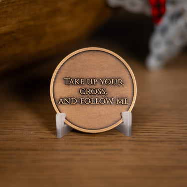 A round bronze-colored metal coin plaque with the engraved text 'TAKE UP YOUR CROSS, AND FOLLOW ME', based on Angela Johnson's work Because of Love, displayed on a small clear acrylic stand on a wooden surface.